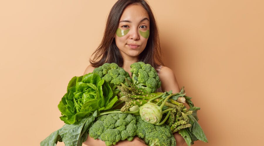 Asian woman with dark hair looks seriously at camera raises eyebrows carries big heap of fresh vegetables applies hydrogel patches under eyes to reduce wrinkles poses against beige background
